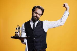 Tired restaurant worker feeling sleepy in studio, yawning and stretching on the job. Butler in suit and tie feeling exhausted and almost falling asleep on camera, poses against yellow background. photo