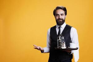 Professional butler serving cup of coffee for clients at restaurant table, carrying drinks on tray for excellent luxury service. Young adult in formal attire acting friendly with customers on camera. photo