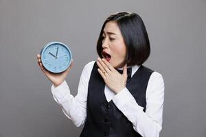 Tired overslept asian waitress in uniform holding circle dial alarm clock and checking time. Restaurant sleepy woman receptionist looking at round retro watch while yawning photo