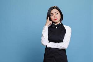 Young asian waitress with pensive facial expression holding finger on head and looking away. Attractive woman receptionist wearing uniform deep in thought posing in studio photo