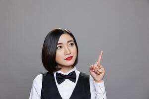Young attractive asian woman receptionist pointing and looking upwards, showcasing direction. Restaurant waitress wearing uniform showing with finger up while posing in studio photo