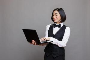 Smiling asian waitress in professional uniform using laptop, checking catering service orders. Cheerful woman receptionist holding portable computer and typing on keyboard photo
