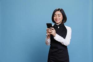 Hotel receptionist holding smartphone and checking message from satisfied customer. Asian waitress with excited face expression reading service positive reviews on mobile phone photo