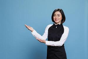 Smiling asian waitress dressed in restaurant uniform showing to side with arms portrait. Receptionist pointing to right direction and looking at camera with joyful facial expression photo