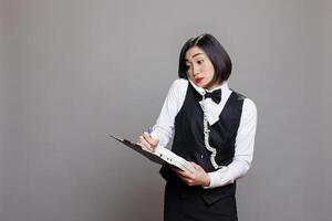 Confused young asian waitress managing conversation on landline phone and writing on clipboard. Restaurant receptionist in uniform maintaining records while chatting with customer on telephone photo