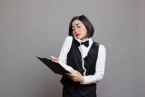 Young asian waitress dressed in uniform writing on a clipboard while talking on landline phone. Restaurant young receptionist chatting with manager on telephone and taking notes photo