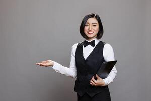 Smiling asian waitress holding digital tablet and welcoming guest with hand portrait. Restaurant receptionist standing with portable device and showing with arm while looking at camera photo