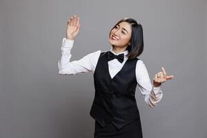 Cheerful asian waitress in uniform laughing and dancing while having fun. Joyful smilng young woman receptionist moving body and hands to music beats while relaxing in studio photo