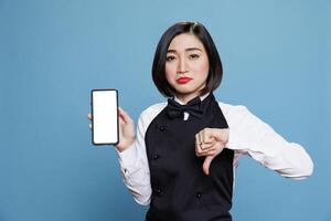 Asian waitress displaying smartphone blank touchscreen mockup and showing thumb down gesture portrait. Disappointed woman receptionist presenting mobile phone empty screen and expressing dissatisfaction photo