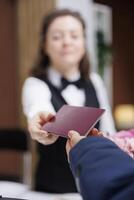 Close-up shot of female concierge returning identity card to traveler wearing winter jacket after check-in process at front desk. Detailed view of passport being grasped by receptionist and tourist. photo
