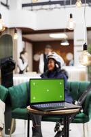 Detailed shot of digital device with isolated copyspace template placed on bench in lounge area of luxury ski mountain resort. Woman in wintersports gear at background of laptop with green screen. photo