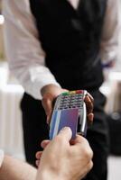 Photo focus of elderly caucasian man tapping his credit card on payment machine held by waiter in lounge area. Detailed view showing a person paying on pos terminal for reservation in hotel lobby.