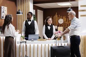 Two guests, elderly couple, arrive at hotel reception for check-in. Receptionists assists senior guests with booking and paperwork, creating welcoming atmosphere. Hotel lobby is lively with tourists. photo