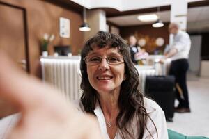 Retired old woman using mobile device to record holiday trip while waiting for check-in. In lounge area, smiling elderly lady snaps photo on her phone while staff aids another client in background.