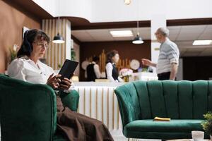 Caucasian elderly lady with headphones, browsing on digital tablet and enjoying vacation. Friendly receptionist assists client with check-in at hotel lobby. Emphasis on hospitality and technology. photo