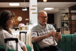 On couch in lounge, senior guy visitor using smartphone to browse the internet. Retired elderly husband utilizing mobile device to make online reservations while sitting on comfortable sofa with wife. photo