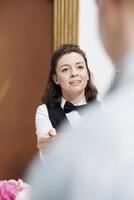 Photo focus on female receptionist helping elderly man at front desk with check in and confirm bookings. Close-up of concierge returning passport to retired senior tourist after verification.