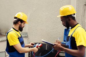Competent mechanic using manifold gauges to read pressure of liquids and gases in hvac system while wireman writes air conditioner inspection and safety check report on clipboard photo