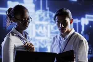African american admin and colleague doing maintenance on artificial intelligence neural networks simulating human thought processes. Diverse experts doing checkup on AI systems using devices photo