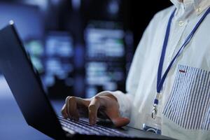 Tech support in data center uses laptop to prevent system overload during peak traffic periods. Hardworking expert in server farm ensuring enough network bandwidth for smooth operations, close up photo
