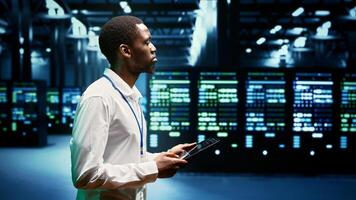 Electrician inspecting server racks delivering web content to customers over the internet. African american wireman using tablet to take note of critical systems, looking for potential errors photo