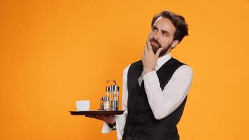 Skilled butler calculating meal prices to give check to clients on camera, serving customers with food or drinks against yellow background. Elegant employee in suit acting thoughtful in studio. photo