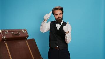 Hotel concierge shows thumbs up in studio, acting positive and optimistic against blue background. Joyful doorman does like and agreement symbol of acceptance, wearing formal attire. photo