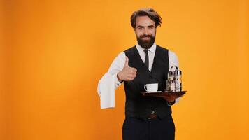 Anccomplished valet raises thumbs up in studio, displays agreement emblem, is enthusiastic on camera. Before serving beverage, young man with a waiter job gestures approbation with like sign. photo