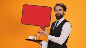 Classy waiter holds speech bubble icon on camera, working to create new advertisement using empty red cardboard. Experienced employee in suit presenting blank carton billboard, serving personnel. photo