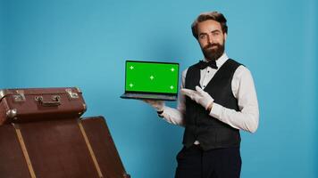 Classy bellhop presents greenscreen on laptop display in studio while he wears gloves next to pile of luggage. Experienced doorman working at luxurious hotel, showing isolated mockup. photo