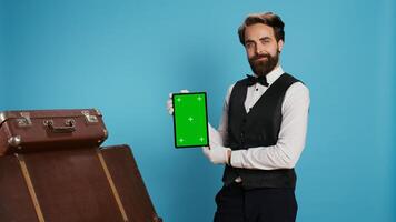 Elegant bellboy holding tablet on camera and presenting blank greenscreen display, symbolizing hotel concierge services with professionalism. Porter in suit and tie pointing at isolated mockup. photo