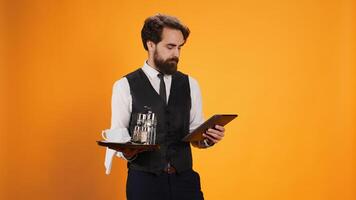 Dedicated staff member calculates check on tablet while working in five star restaurant to serve clients, offer outstanding opulent service. Butler carrying dinner platter and modern gadget. photo