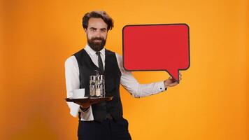 Catering worker presents speech bubble with blank banner space on red icon cutout in studio, indicating advertisement with empty cardboard isolated copyspace. Young butler with food tray. photo