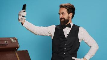 In studio, doorman in uniform takes photos while using smartphone for selfies and acting silly against blue background. Skilled hotel concierge does pictures with his gadget while on the job.