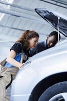 Repairman and coworker collaborating on servicing broken vehicle, checking for faulty distributor. Trained mechanics in auto repair shop working together on fixing car, discussing best options photo