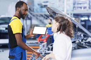 Mechanic in car service uses mockup tablet to order new engine for malfunctioning vehicle. Adept garage workspace expert shows woman needed parts replacement on isolated screen device photo