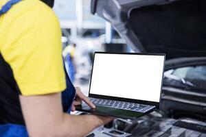 Seasoned mechanic in garage using mockup laptop to follow checklist while doing maintenance on car. Employee in auto repair shop does checkup on vehicle helped by isolated screen device photo