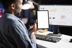 Young man sits at his desk utilizing tablet with empty screen before reviewing fiscal information on computer. At home, african american man handles smartphone with white display layout. photo