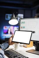Young employee checks tablet with white screen at home, calculating finances and sales trend on computer. Advisor uses pc and device with isolated empty layout, monthly statistics. photo