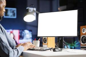 African american man checks white screen on computer, teleworking and using blank display on monitor. Male employee does remote work at home with empty chromakey design at desk. photo