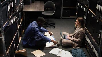 African american policeman reading criminal records files on office floor, trying to uncover details and clues. Multiethnic detectives studying case information, searching for forensic evidence. photo