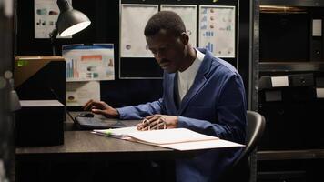Private investigator seen in dimly lit office meticulously analyzing papers and evidence gathered during research. With laptop open, policeman cross-references information and does background check. photo