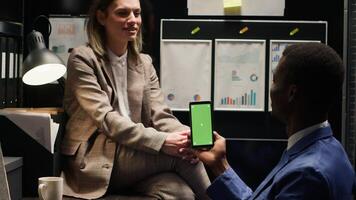 African american investigator conducts inquiry with caucasian officer while utilising chromakey greenscreen display on phone. Detectives in office, examines an isolated mockup template with copyspace. photo