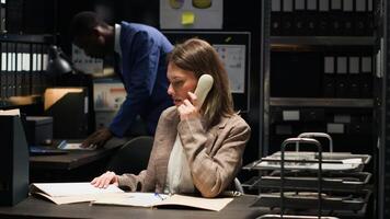 Caucasian private detective answers call to find out about forensic evidence report and analyze classified records. Female investigator examines files and uses landline to confirm confidential clues. photo