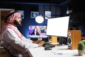 Islamic guy sits at his home office table, his desktop computer monitor displaying a blank white screen. A dedicated Muslim man is utilizing a computer with an isolated chromakey mockup template. photo