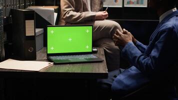 Modern laptop with blank green screen positioned on desk in police evidence room. In the office, wireless computer displays isolated chromakey copyspace template while officers talk in background. photo