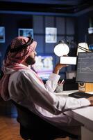 Side-view shot Middle Eastern man in traditional attire sitting at an office desk, using a desktop computer. They hold a mobile phone with a blank chromakey mockup template, offering space for advertising. photo