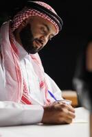 The image portrays an attentive Middle Eastern guy in traditional clothing working at a table with a notepad. An Islamic man is shown writing research at his desk at home in a close-up shot. photo