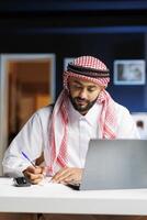 An Arab businessman, absorbed in work, diligently browses digital information on office desk laptop. He takes notes while conducting internet research, demonstrating his professionalism and attention. photo