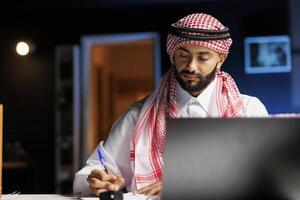 Male Arab entrepreneur working at his modern desk, browsing the internet and writing in his notebook. Image shows a young Muslim man using a pen, book and a minicomputer on the table. photo